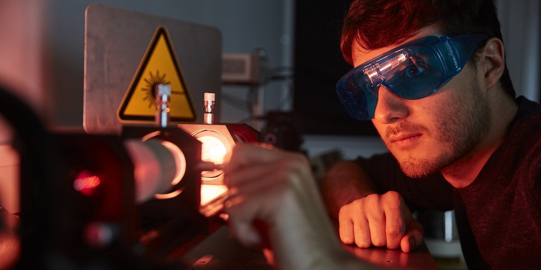 Das Bild zeigt einen Studenten im abgedunkelten Laserlabor. Er hat eine Schutzbrille auf und justiert eine Linse im Strahlengang des Lasers.