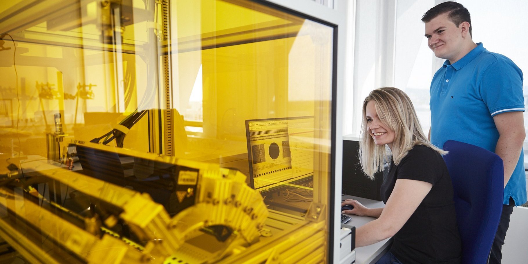 In the foreground you can see a machine in a protective housing made of yellow-colored Plexiglas. A thick cable is led outside to a computer. A student sits there and operates the mouse while she and a student observe whether the machine reacts appropriately to the commands.