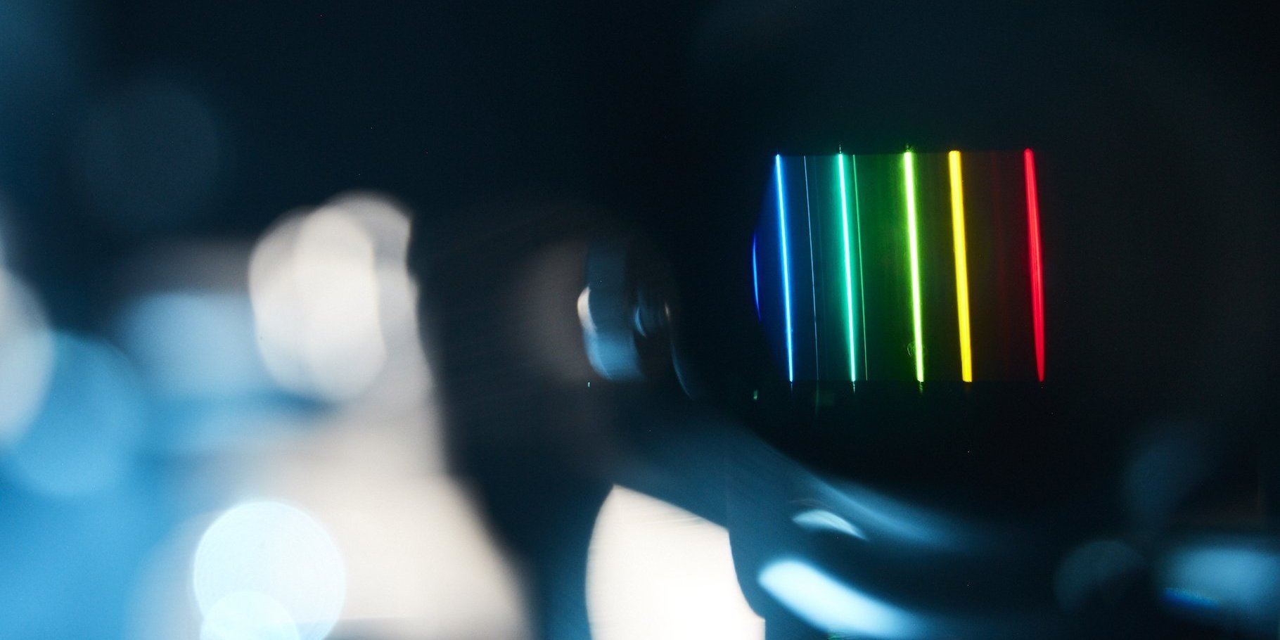 Im Hintergrund sind scharf Spektrallinien zu sehen, von tiefem Blau über Türkis und Hellgrün bis zu strahendem Gelb und kräftigem Rot. Die Geräte im Vordergrund sind aufgrund der Kameraeinstellungen nur sehr unscharf wahrzunehmen und nicht klar zu erkennen.