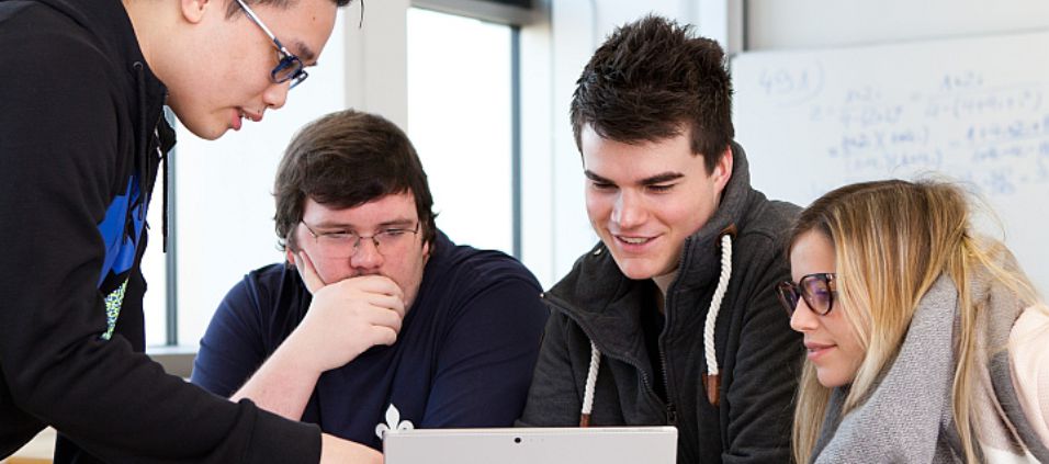 Zwei Studenten und eine Studentin sitzen vor einem Notebook. Ein vierter Student steht dabei. Er zeigt dabei erklärend auf etwas auf dem Bildschirm während die anderen drei aufmerksam zuhören.