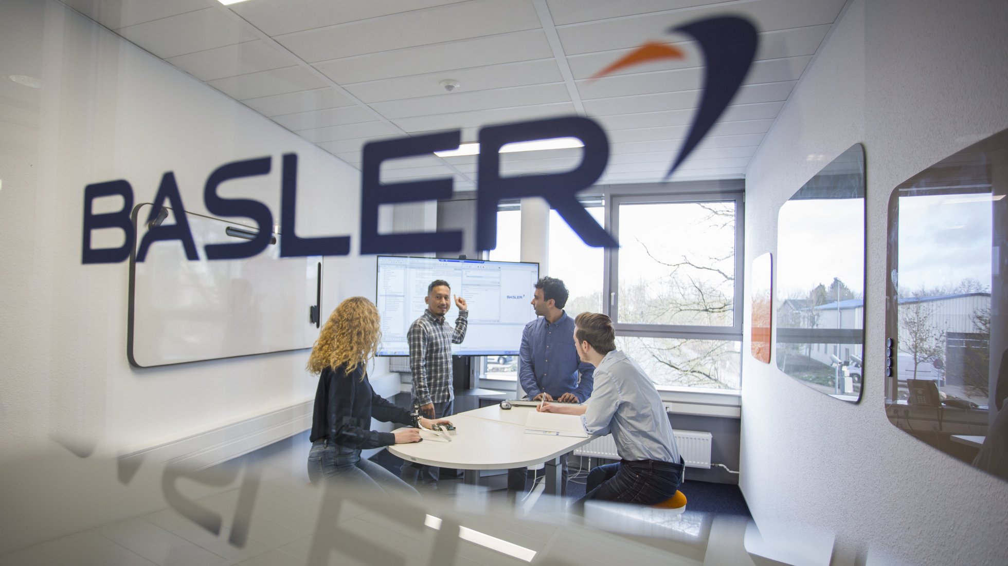 View through a glass front into a meeting room at Balser. A young man is explaining something on a large screen while three other people listen.