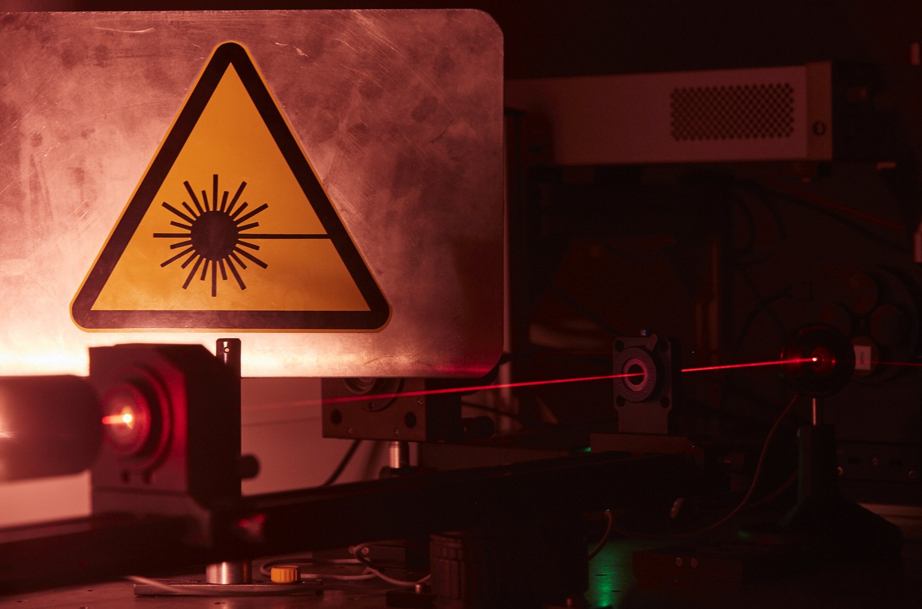 Das Bild zeigt ein Detail eines Aufbaus im abgedunkelten Laserlabor. Es ist gut der rote Laserstrahl zu erkennen. Im Hintergrund hängt groß das dreieckige gelbe Schild, das vor dem Laserlicht warnt.