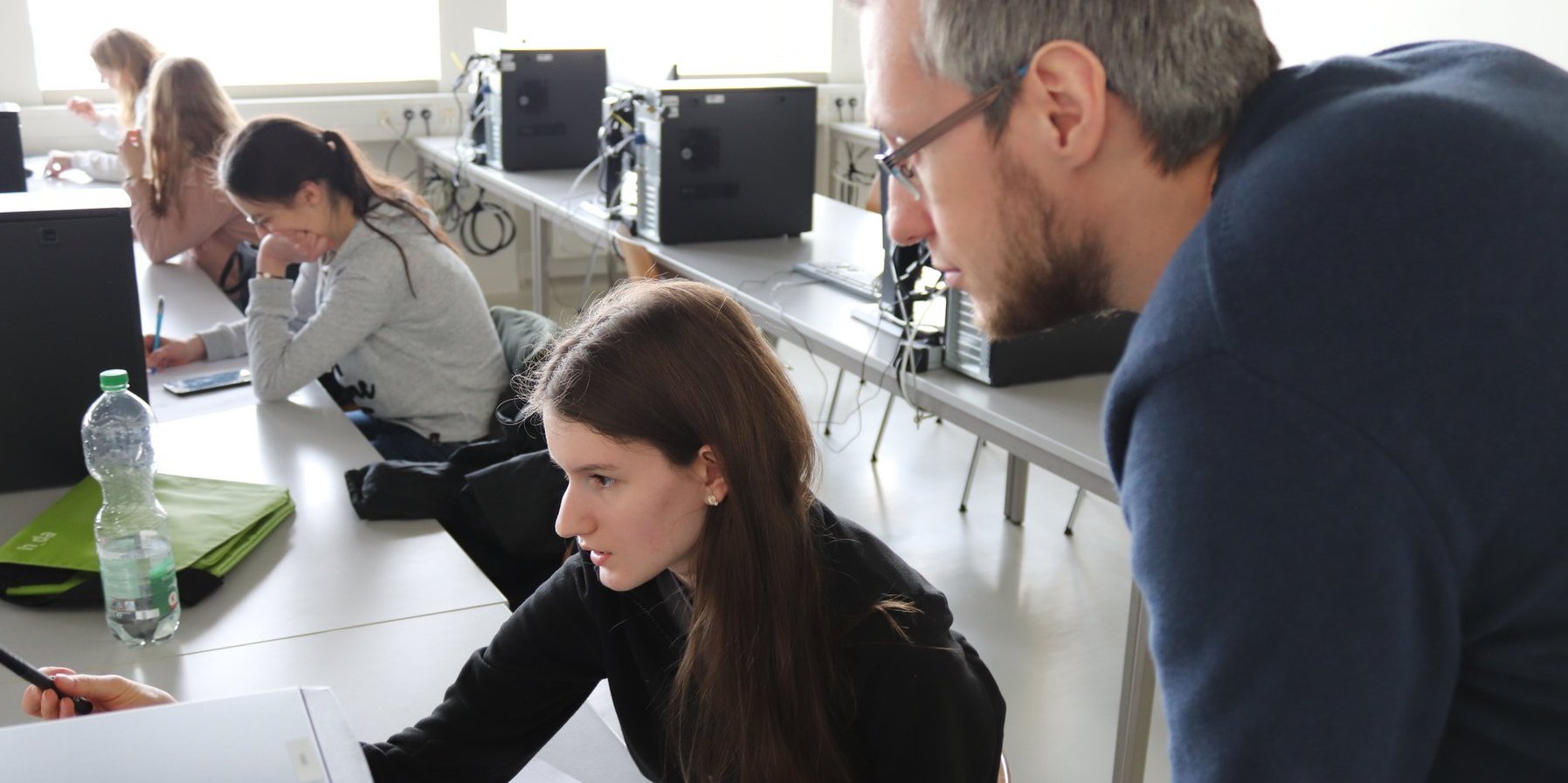 Ein Laboringenieur hört sich die Frage an, die eine Teilnehmerin stellt und dabei auf den Bildschrim deutet. Im Hintergrund sind weitere junge Frauen zu sehen, die ebenfalls vor den Bildschirmen sitzen oder sich Notizen machen.