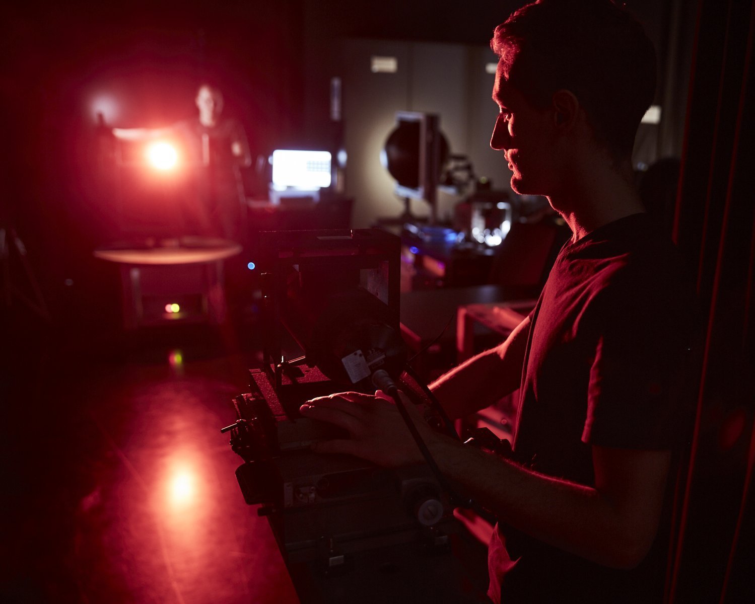 We look into a darkened laboratory. In the background you can dimly make out a student aiming a lamp. The light source shines directly into the camera and the scattered light illuminates the room in a faint red. In the foreground is a second student pointing a detector at the light source. 
