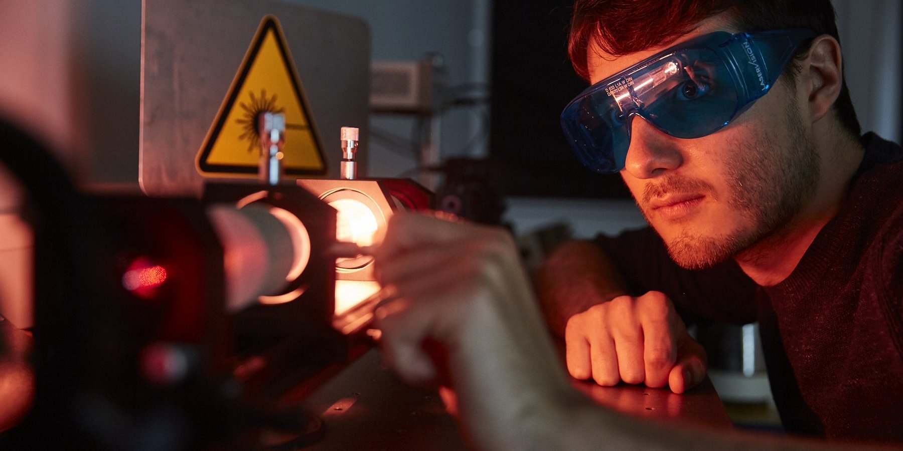 Das Bild zeigt einen Studenten im abgedunkelten Laserlabor. Er hat eine Schutzbrille auf und justiert eine Linse im Strahlengang des Lasers.