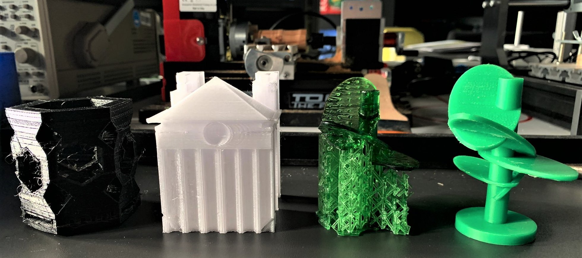 Das Foto zeigt vier Objekte, die im Rahmen des Schulpraktikums mit 3D-Druckern hergestellt wurden: einen achteckigen Stifthalter aus schwarzem Kunststoff, ein Gebäude mit einem Säulenportal aus weißem Kunststoff und aus grünem Kunststoff zwei futuristische, sehr filigrane Bäume.