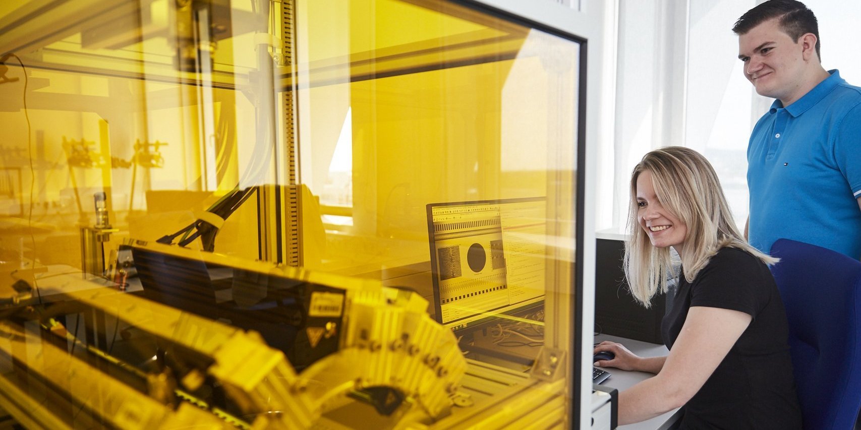 Im Vordergrund sieht man in einem Schutzgehäuse aus gelb eingefärbtem Plexiglas eine Maschine. Ein dicker Kabelstrang wird nach außem zu einem Rechner geführt. Dort sitzt eine Studentin und bedient die Mouse, während sie gemeinsam mit einem Studenten beobachtet, ob die Maschine entsprechend auf die Befehle reagiert.
