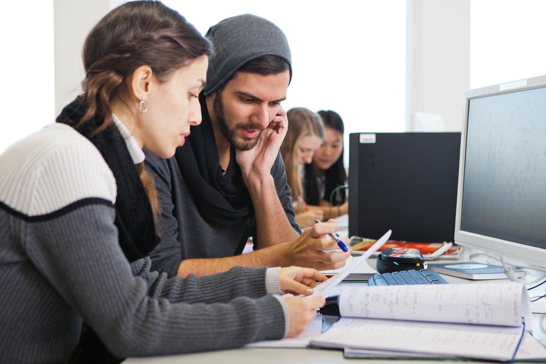 Eine junge Frau und ein junger Mann sitzen vor einem Bildschirm und diskutieren. Im Hintergrund sind zwei weitere Studentinnen zu erkennen, die sich ebenfalls unterhalten.