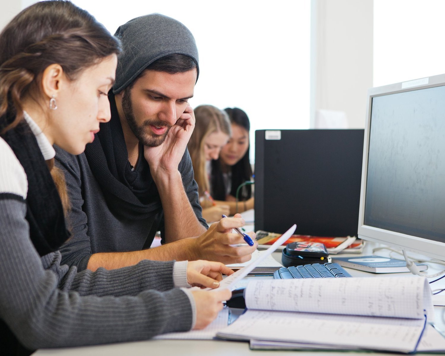 Eine junge Frau und ein junger Mann sitzen vor einem Bildschirm und diskutieren. Im Hintergrund sind zwei weitere Studentinnen zu erkennen, die sich ebenfalls unterhalten.